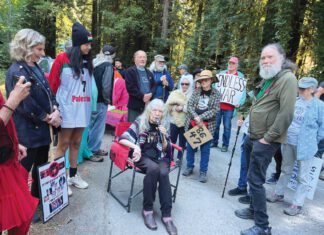 Bohemian Groove, Protest ‘Worth the Effort’ Woman seated on chair speaking outdoors to a gathered crowd.