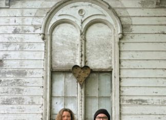 Around the Sun and Up the Wall Two musicians standing with an acoustic guitar in front of a weathered white building