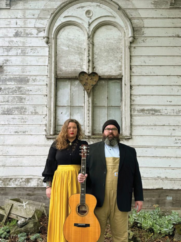 Two musicians standing with an acoustic guitar in front of a weathered white building