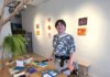 Artist standing beside a table covered with paintings and art supplies in a gallery