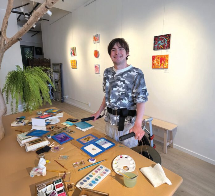 Artist standing beside a table covered with paintings and art supplies in a gallery