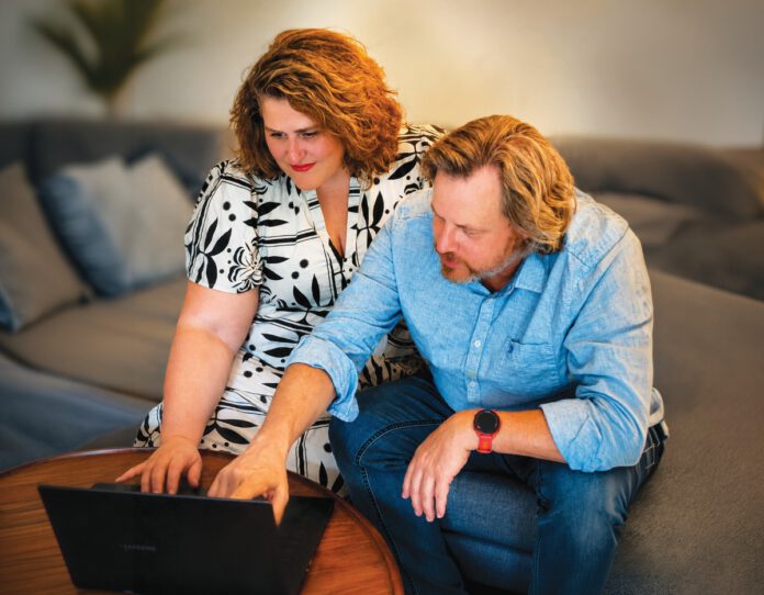 A woman and man sit together on a couch, looking at a laptop
