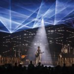 Third Eye Blind, a five-member rock band, performs onstage beneath blue laser lights and a central white spotlight as an audience watches.