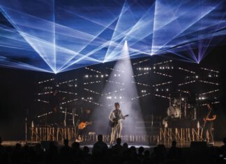 Third Eye Blind, a five-member rock band, performs onstage beneath blue laser lights and a central white spotlight as an audience watches.