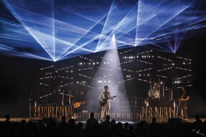Third Eye Blind, a five-member rock band, performs onstage beneath blue laser lights and a central white spotlight as an audience watches.