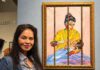 A woman in a denim jacket stands beside a framed, brightly colored painting of a seated figure wrapped in a yellow garment.
