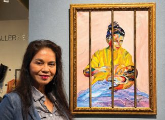A woman in a denim jacket stands beside a framed, brightly colored painting of a seated figure wrapped in a yellow garment.