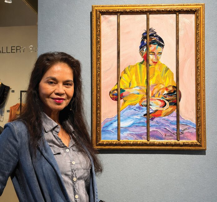 A woman in a denim jacket stands beside a framed, brightly colored painting of a seated figure wrapped in a yellow garment.