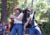 Two musicians in cowboy hats and sunglasses play electric guitars back to back on an outdoor stage, with trees behind them.