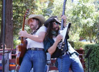 Two musicians in cowboy hats and sunglasses play electric guitars back to back on an outdoor stage, with trees behind them.