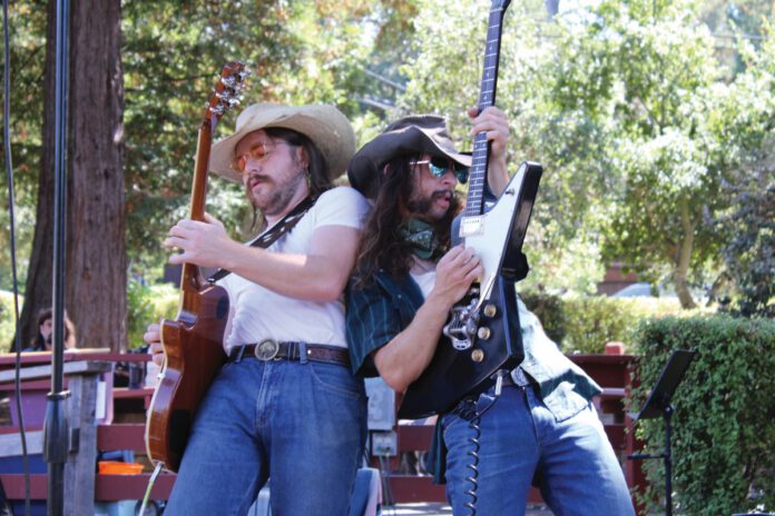 Two musicians in cowboy hats and sunglasses play electric guitars back to back on an outdoor stage, with trees behind them.