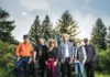Six musicians stand together outdoors near pine trees at dusk.