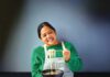 A smiling woman in a green sweater and apron lifts noodles with chopsticks over a bowl of soup.