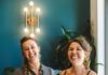 Two smiling women hold fancy frosted cakes on glass pedestal stands.