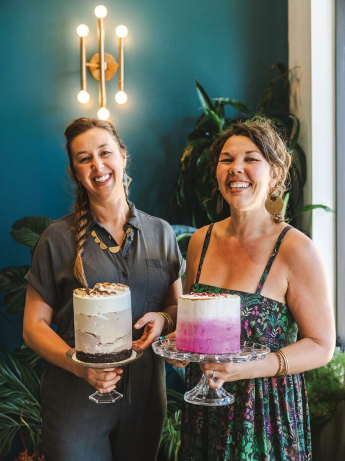 Two smiling women hold fancy frosted cakes on glass pedestal stands.
