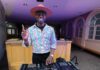 A DJ in a wide-brimmed pink hat and heart-patterned jacket extends his index finger up while working a turntable in a room with purple light.