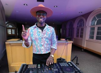 A DJ in a wide-brimmed pink hat and heart-patterned jacket extends his index finger up while working a turntable in a room with purple light.