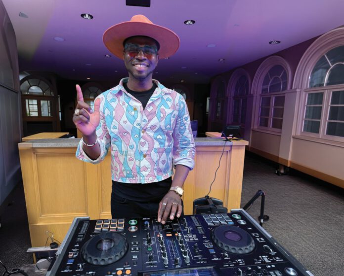 A DJ in a wide-brimmed pink hat and heart-patterned jacket extends his index finger up while working a turntable in a room with purple light.