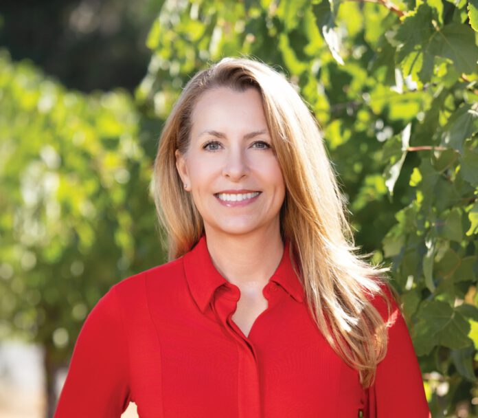 A woman with long blonde hair and a red button-down shirt smiles in front of green grapevines.