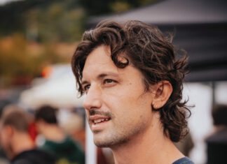 A man with curly brown hair and a mustache wears a navy t-shirt.