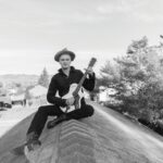 A black-and-white photo of a man in a wide-brimmed hat and dark clothing sitting cross-legged on a rooftop peak, holding a guitar.
