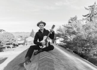 A black-and-white photo of a man in a wide-brimmed hat and dark clothing sitting cross-legged on a rooftop peak, holding a guitar.