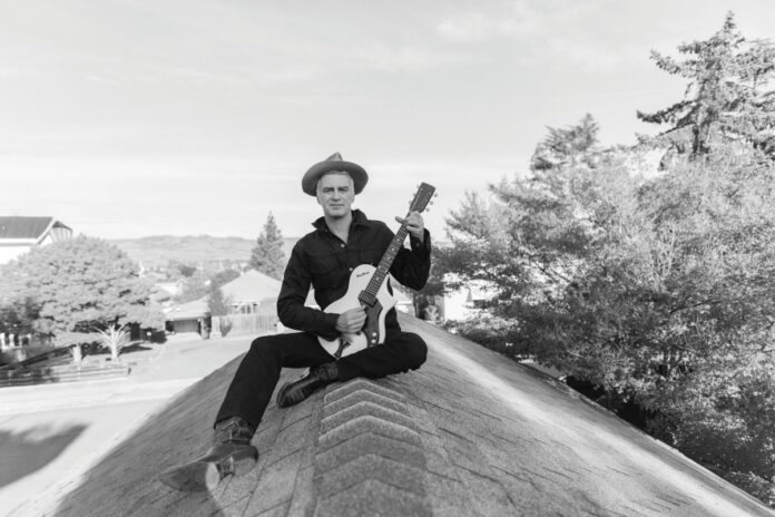 A black-and-white photo of a man in a wide-brimmed hat and dark clothing sitting cross-legged on a rooftop peak, holding a guitar.