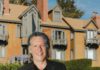 A smiling man in a blazer stands on a lawn in front of a large shingled house with balconies.