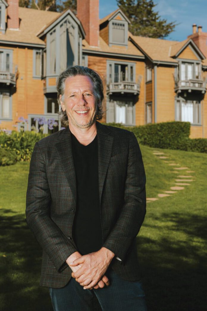 A smiling man in a blazer stands on a lawn in front of a large shingled house with balconies.
