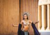 A cosplayer in an ornate gold headdress and jeweled costume poses dramatically in front of stone columns.