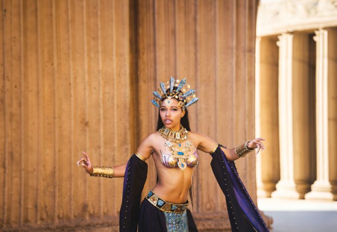 A cosplayer in an ornate gold headdress and jeweled costume poses dramatically in front of stone columns.