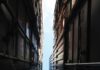 A narrow gap between tall stacked shipping containers opens to a sliver of blue sky.