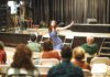 A woman in a striped dress performs, arms outstretched, while an audience watches from folding chairs.