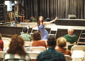 A woman in a striped dress performs, arms outstretched, while an audience watches from folding chairs.