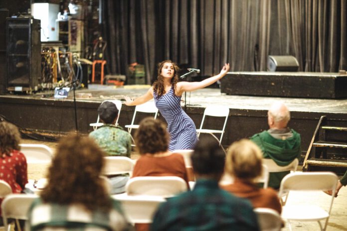 A woman in a striped dress performs, arms outstretched, while an audience watches from folding chairs.