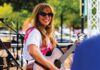 A woman with long hair and sunglasses smiles while playing an acoustic guitar on an outdoor stage.