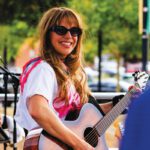 Last Krush of Summer: Danny Sorentino and Anna Jae Rock Vintage Space A woman with long hair and sunglasses smiles while playing an acoustic guitar on an outdoor stage.
