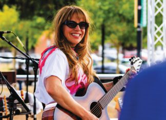 A woman with long hair and sunglasses smiles while playing an acoustic guitar on an outdoor stage.