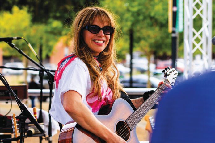 A woman with long hair and sunglasses smiles while playing an acoustic guitar on an outdoor stage.