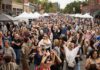 A crowd of people with arms raised dance at an outdoor festival with vendor tents in the background.