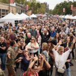 Tunes & Tracks: Railroad Square Music Fest Rocks for Free A crowd of people with arms raised dance at an outdoor festival with vendor tents in the background.