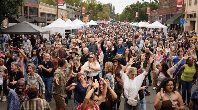 A crowd of people with arms raised dance at an outdoor festival with vendor tents in the background.