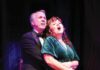 A man in a tuxedo stands behind a woman in a green dress, wrapping his arms around her waist as they both sing.