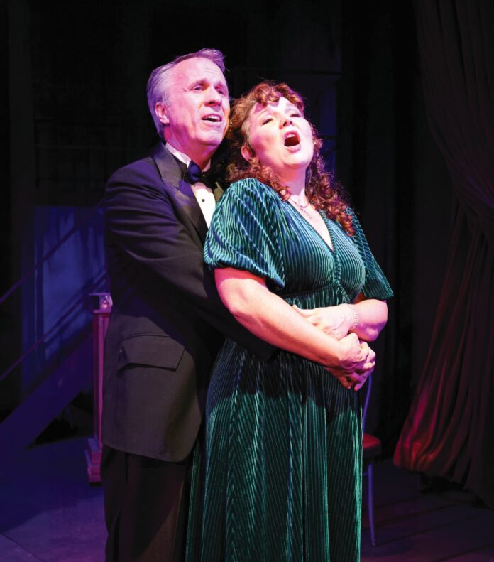 A man in a tuxedo stands behind a woman in a green dress, wrapping his arms around her waist as they both sing.