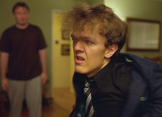 A young man in a blue jacket and tie looks uncomfortable, with another man in a brown shirt standing behind him in a living room.