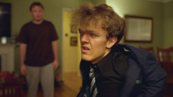 A young man in a blue jacket and tie looks uncomfortable, with another man in a brown shirt standing behind him in a living room.