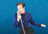 American Idolatry: Musical Satire of Elvis Hysteria at Cinnabar A man in a shiny blue shirt and black tie sings into a vintage microphone against a blue background with "Bye Bye Birdie" written on it many times.