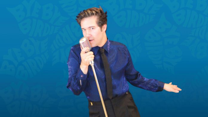 A man in a shiny blue shirt and black tie sings into a vintage microphone against a blue background with 