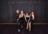 Four performers in glittering black outfits pose together against a dark, ornate wallpapered backdrop.