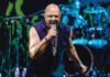 A bald singer in a sleeveless purple shirt sings into a microphone on a blue-lit stage with drums behind him.